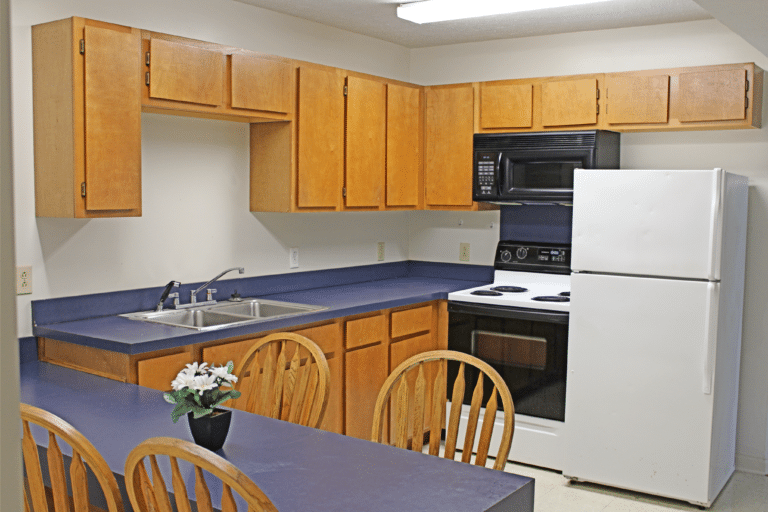 The kitchen inside of on apartment in Trustee Hall. There is a refrigerator, stove, microwave, sink, and table.